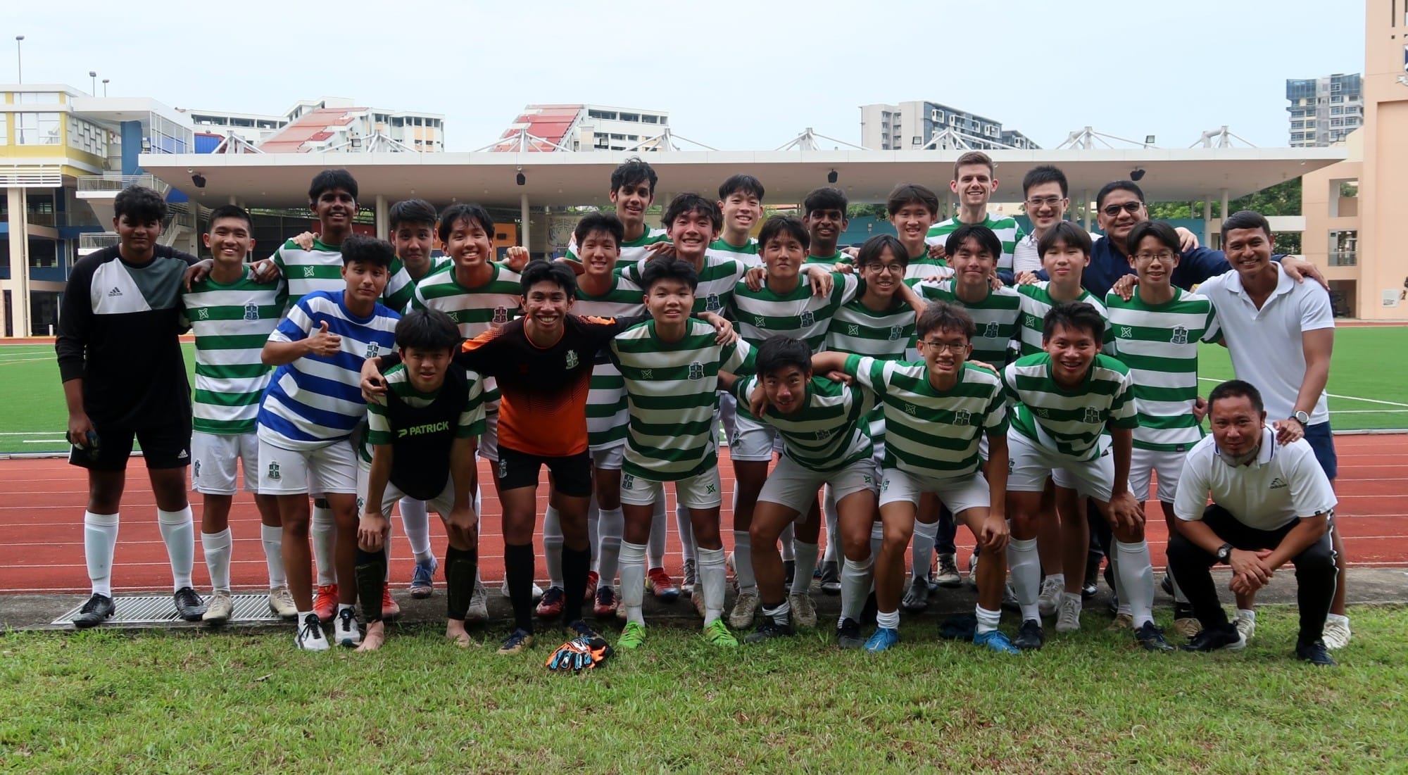 A school football team in green and white hooped jerseys poses together on the field with coaches, after a match.